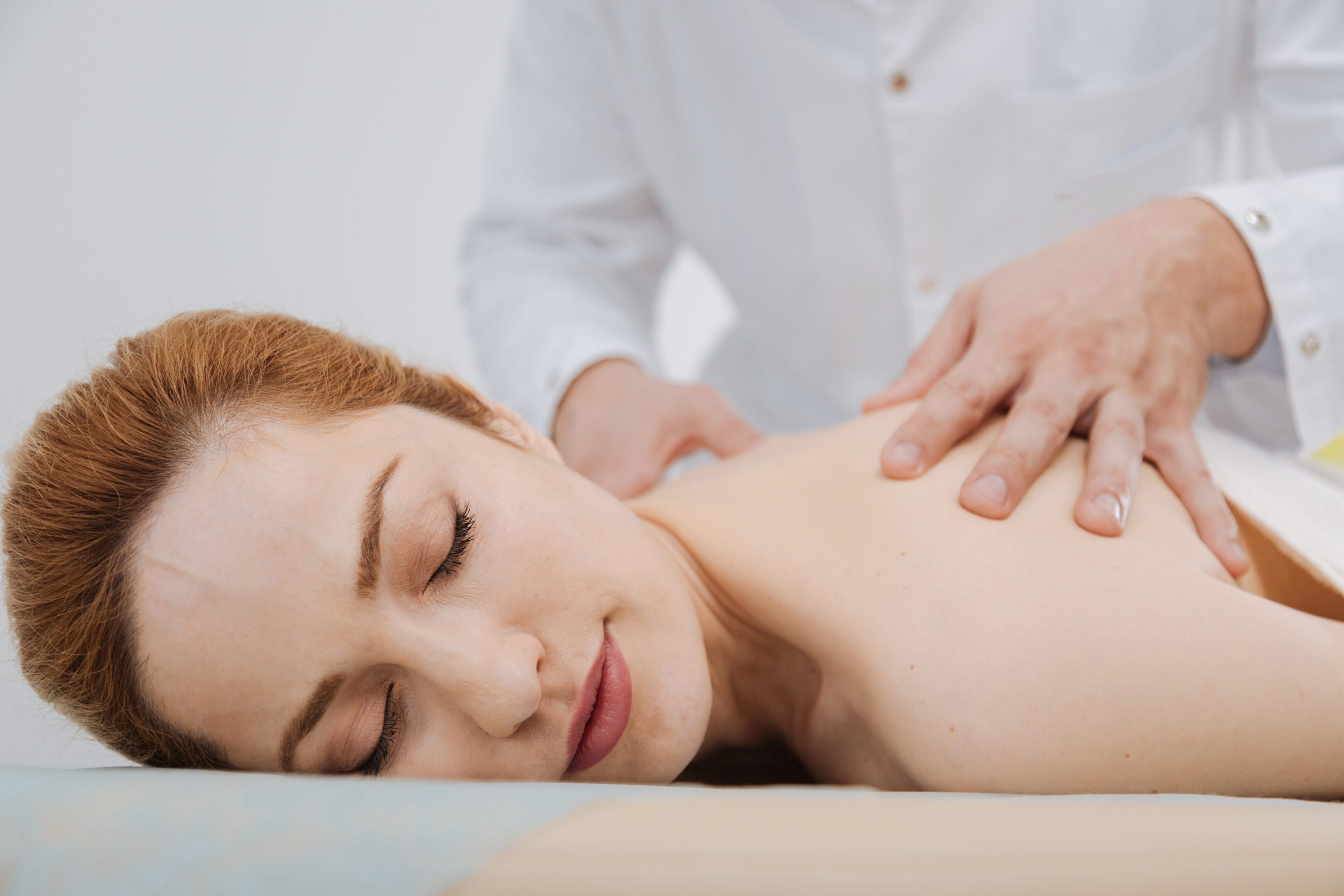 Haling touch. Pretty weary young woman lying on bed and holding her eyes closed while the doctor massaging the muscles on her back